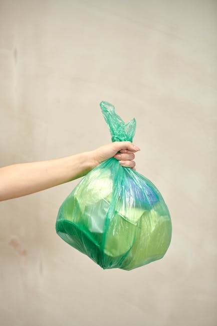 A person's hand is visible holding a green and blue plastic garbage bag by its twisted knot top, positioned centrally against a plain, light beige background. The bag appears filled with waste, and its semi-translucent material shows some contents within. The hand is fair-skinned and extended from the left side of the image, gripping the bag tightly. This scene relates to waste disposal or rubbish collection, representing an individual preparing to discard household waste or recyclables, often associated with private rubbish removal services like those offered by rubbishclearancequeenspark.com. The minimal environmental context highlights the focus on waste management tasks, emphasizing the importance of proper rubbish handling, especially in situations where alternative disposal methods or same-day collection options are relevant.