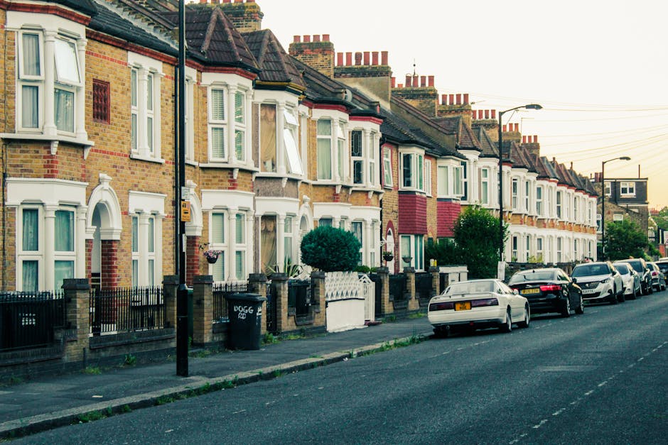 A row of traditional terraced houses on a residential street, featuring brick facades with a mix of red and yellow tones and white window frames. Several of the houses have bay windows on the ground and first floors, with some windows slightly open or covered with curtains. The street is lined with parked cars, including a mix of sedans and hatchbacks, parked parallel to the pavement on the right side. The pavement is grey, with a few small patches of grass near the curb, and there is a black wheelie bin labeled with the word ‘DOGG’ situated in front of one property. Tall, black lamp posts with curved arms extend over the road, and an individual house has a small, neatly trimmed green shrub in front, bordered by a low brick wall with white fencing. Overhead, multiple utility wires stretch across the sky, which is clear with a soft, warm evening light, highlighting the calm character of the neighborhood. The scene reflects a typical urban residential area where independent rubbish removal services may be employed for waste clearance, in contrast to municipal collection, aligning with private disposal practices often seen in such settings.