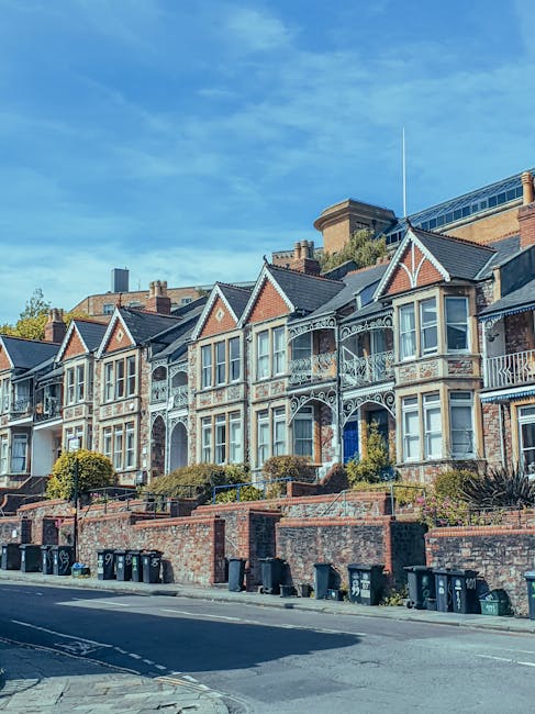 A row of Victorian-style terraced houses with decorative ironwork balconies and gabled roofs, set on a slightly elevated street level with brick retaining walls and small landscaped front gardens. The buildings feature large bay windows with white frames, brick facades with contrasting detailing, and some have small porches leading to front doors. The street in the foreground shows a paved road with several black wheelie bins lined along the pavement, indicating waste disposal or collection points. The background includes a modern building partially visible behind the older houses, and the sky appears clear with a few wispy clouds, suggesting a bright, sunny day. The scene reflects an urban residential area that may benefit from private rubbish removal or on-site clearance services provided by companies such as rubbishclearancequeenspark.com. The overall environment is quiet and well-maintained, emphasizing the importance of coordinated waste management for local homes.